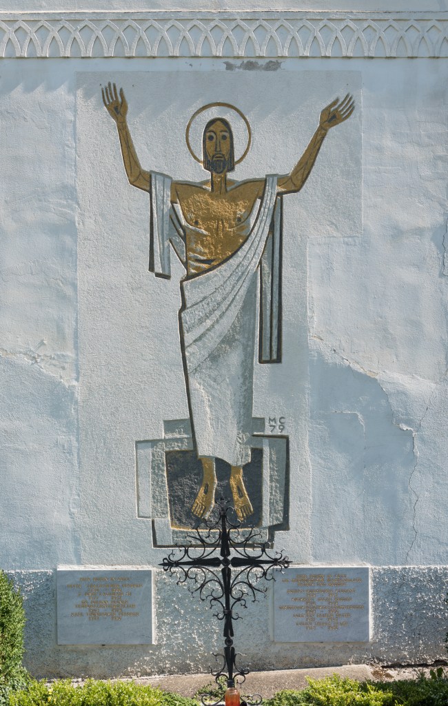 A painting of a full figure of Christ, arms raised with brown-gold skin, on a white wall.